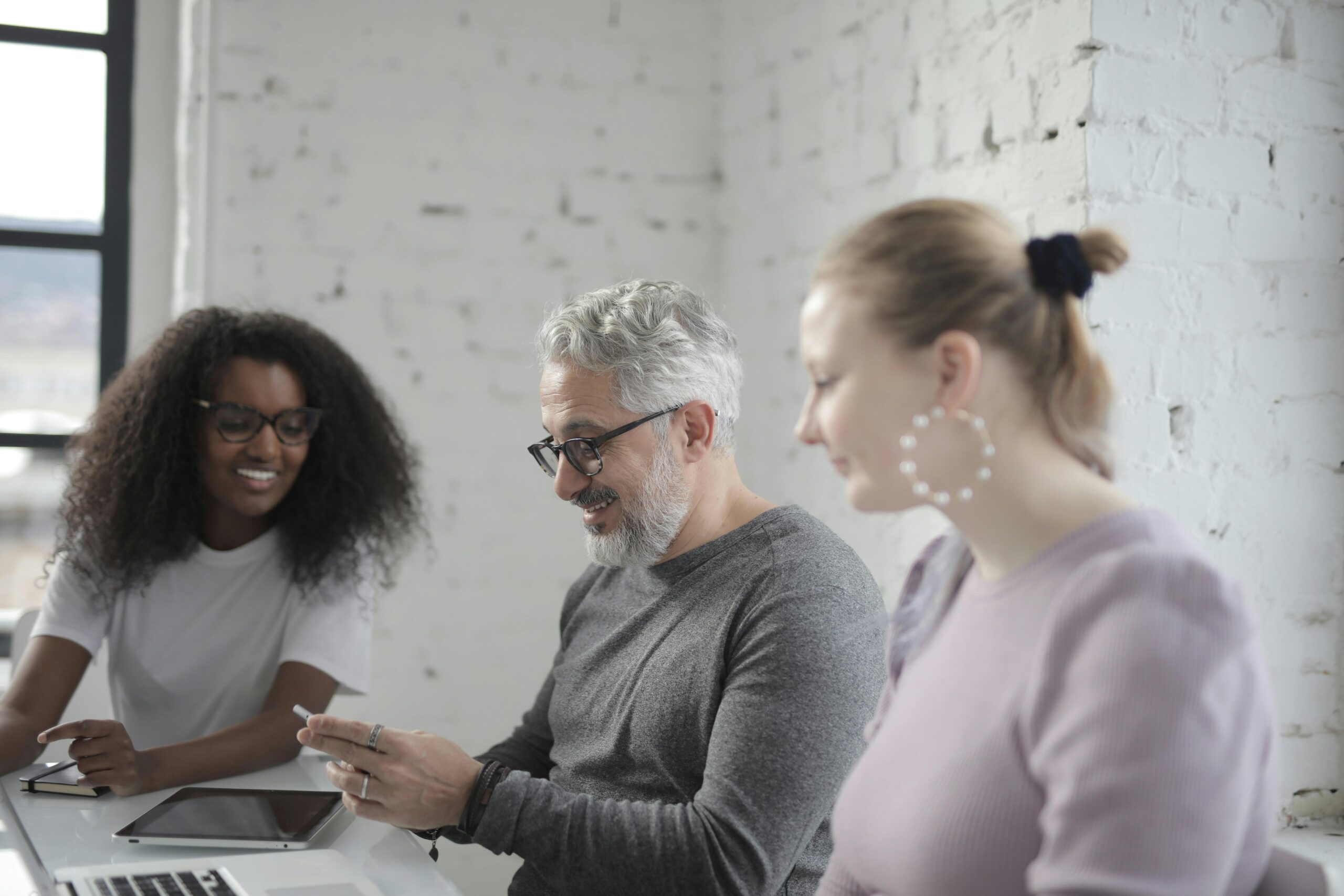 Positive man wearing casual clothes and eyeglasses sitting at table with laptop and tablet and browsing smartphone with coworkers in office